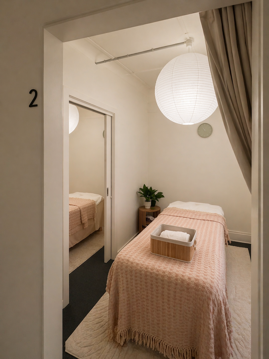Single treatment room with pink throw, paper-lantern pendant and an open door revealing the second room behind